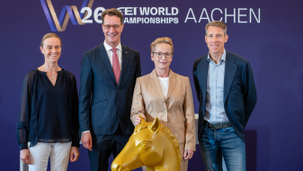 Ministerpräsident Wüst und drei Pferdesport-Athletinnen und -Athleten vor einem Banner der Reit-WM in Aachen, vor ihnen ein goldfarbenes Schachfigur-Pferd