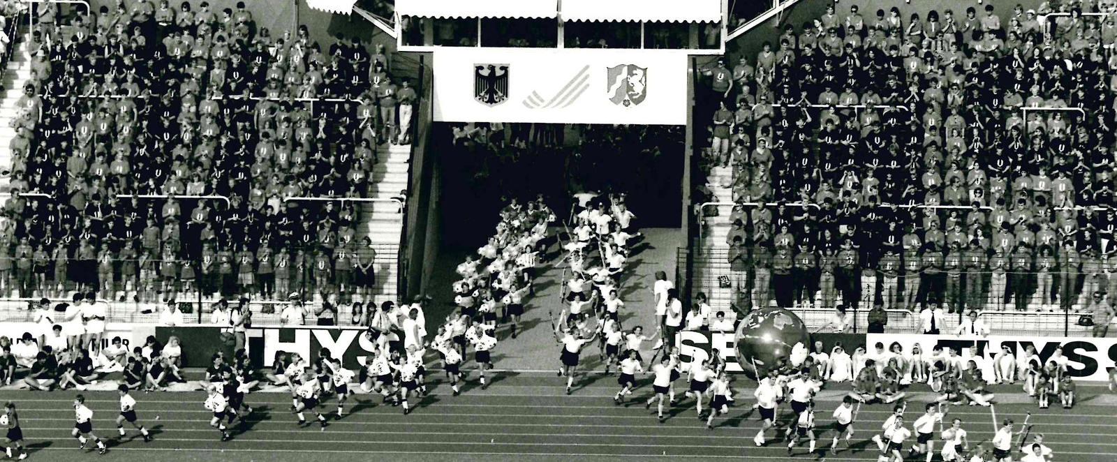 Das schwarz-weiß Foto zeigt Sportler, die zur Universiade vor vollbesetzten Tribühnen in ein Stadion einlaufen.