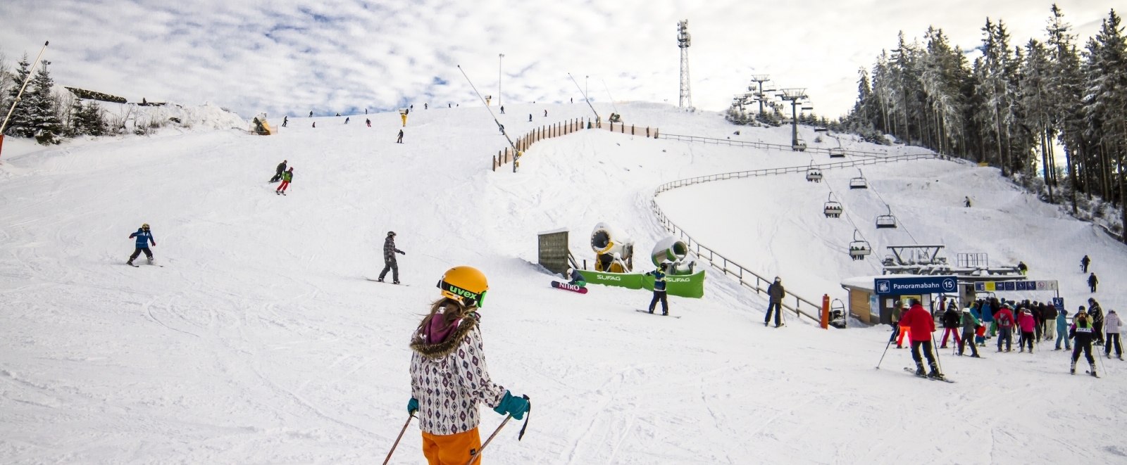 Auf dem Foto sieht man mehrere Skifahrer auf einer schneebedeckten Piste. Rechts sieht man einen Lift und am Rand einen Tannenwald.