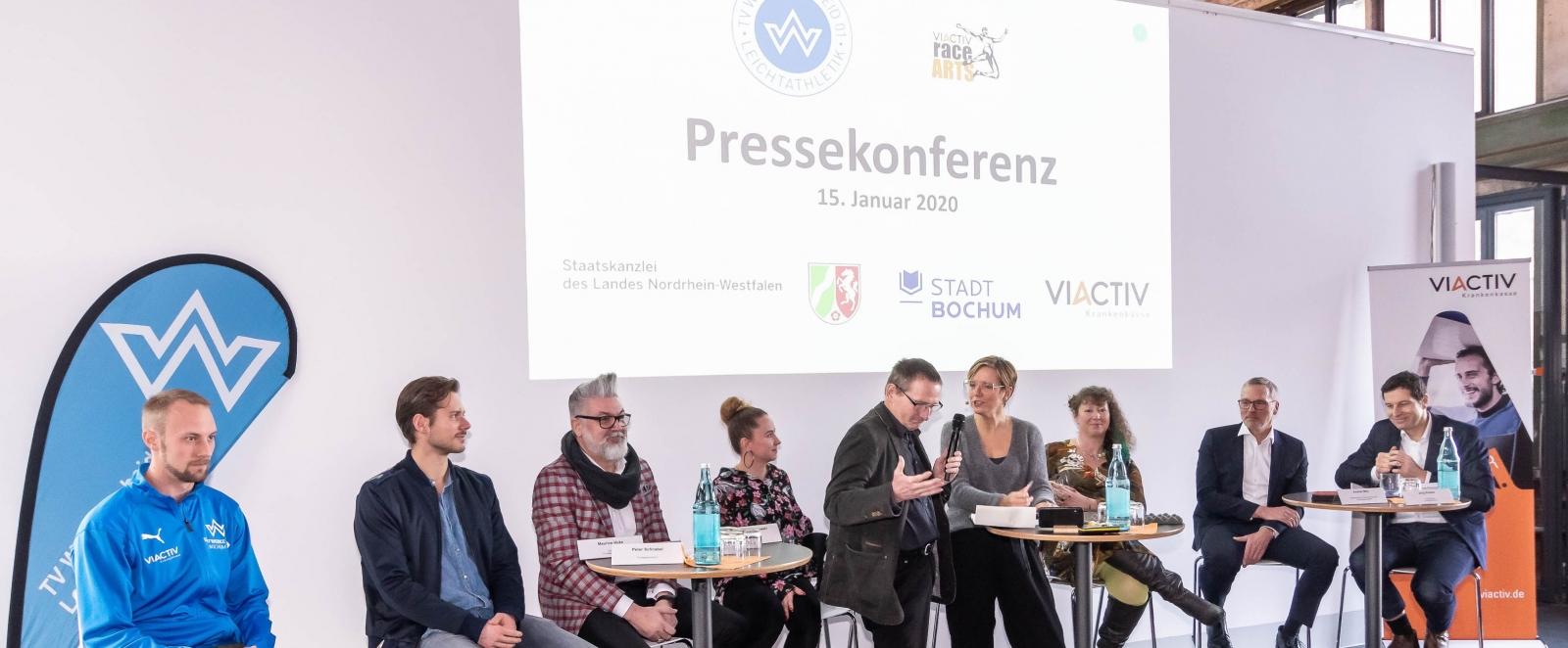 Staatssekretärin Andrea Milz sitzt mit Teilnehmern der Pressekonferenz auf Hockern an Rundtischen. Hinter ihnen eine weiße Rückwand mit einer Präsentation.