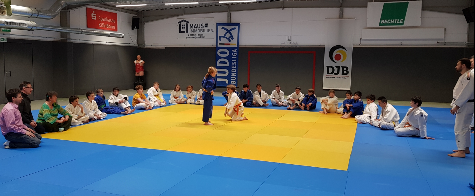 Zwei jugendliche Judokas demonstrieren Selbstverteidigung auf einer gelben, blau-umrandeten Matte. Auf dem breiten blauen Rand sitzen im Halbkreis  weitere Judokas. Rechts am Bildrand steht der Trainer.