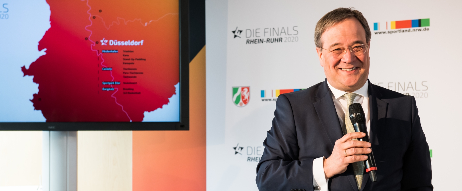 MInisterpräsident Laschet steht vor einer weißen Plakatwand und hält in seiner rechten Hand ein Mikrofon, während er lächelnd leicht nach rechts schaut.