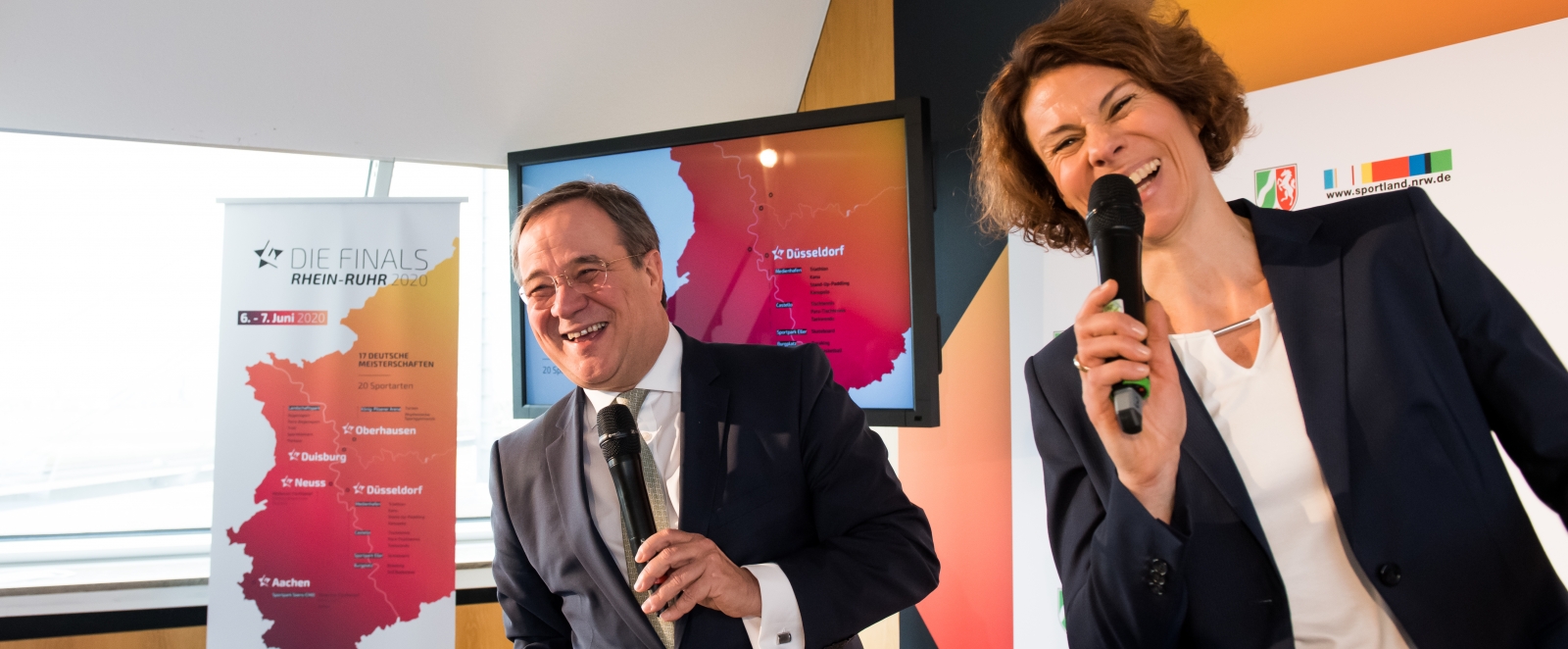 Ministerpräsident Laschet und eine Moderatorin stehen nebeneinander leicht nach vorne links gebeugt und jeder hält ein Mikrofon in der Hand. Vor ihnen steht ein Turngerät namens Pferd.