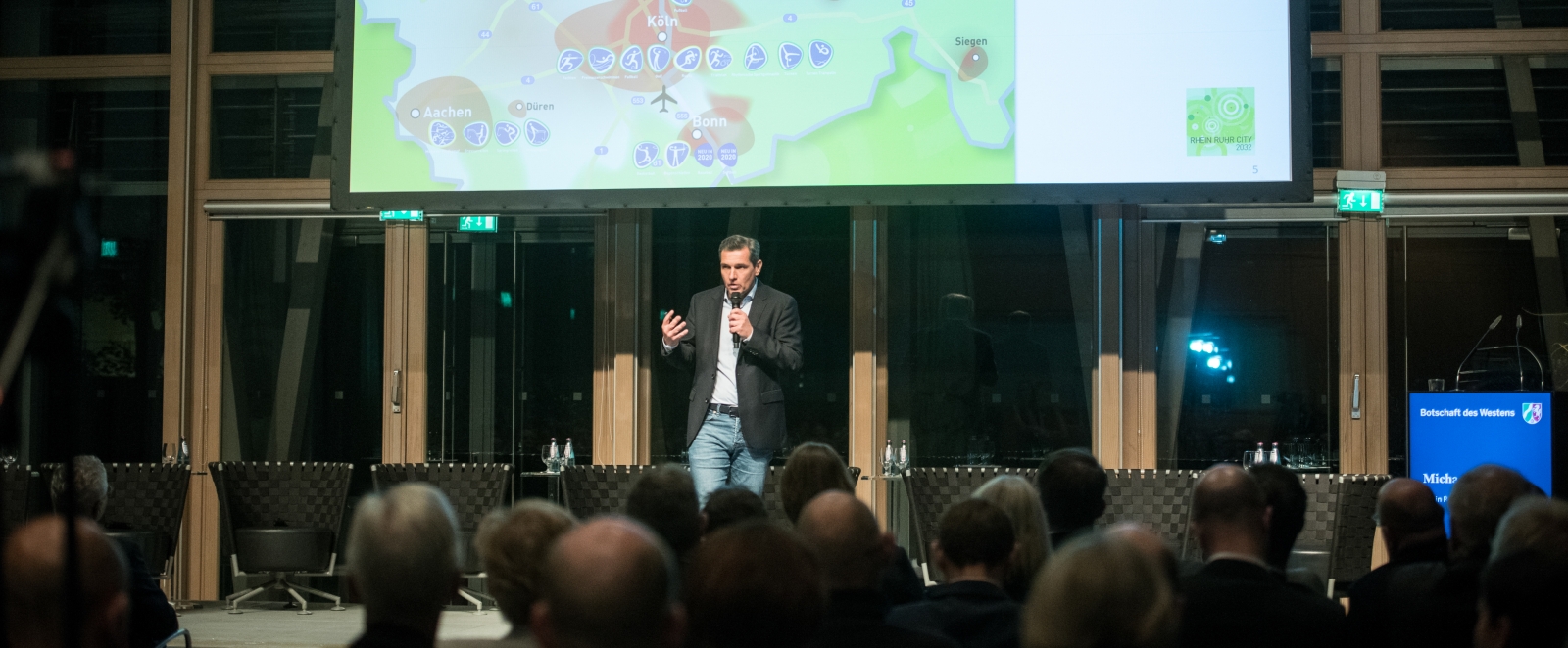 Ein Mann hält eine Rede vor einem großen Publikum in einem Saal. Vorne oben hängt eine Leinwand mit einer Präsentation auf der sieht man eine graphische Darstellung mit den Umrissen von NRW.