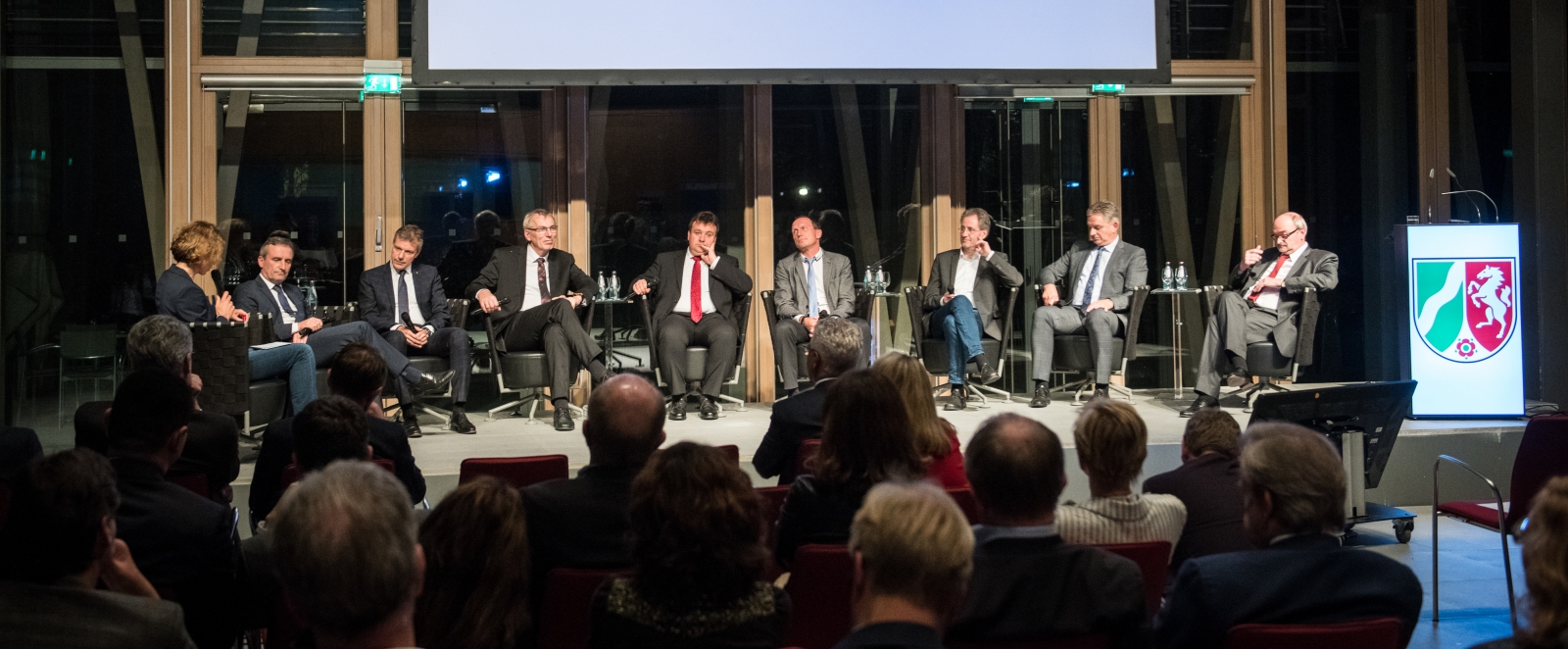 Auf einer Bühne in einem großen Saal sitzen 9 Personen im Halbkreis in einer Diskussionsrunde.