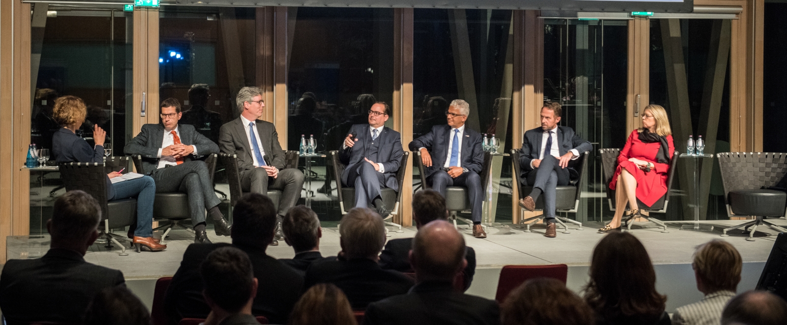 Auf einer Bühne in einem großen Saal sitzen 7 Personen im Halbkreis in einer Diskussionsrunde.