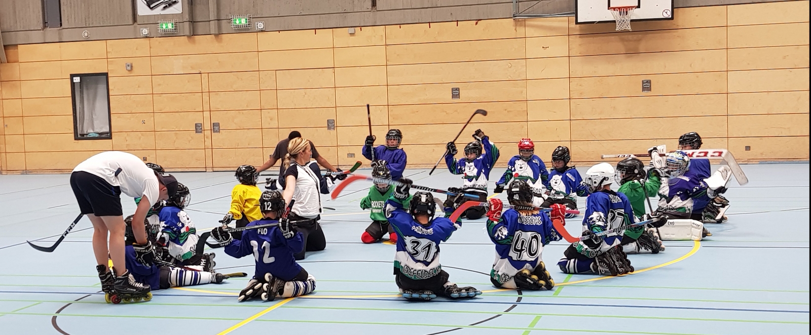 Eine Kinder-Hockeyspielergruppe von Mädchen und Jungen  mit einer Trainerin und einem Trainer in einer Turnhalle.