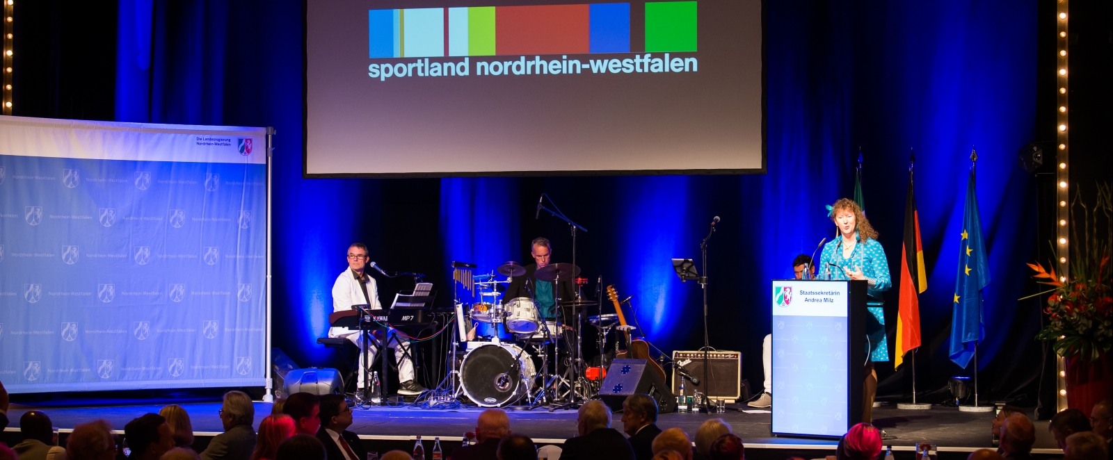 Staatssekretärin Andrea Milz am Rednerpult rechtsseitg auf einer Bühne. Links sitzt eine Musikband. Vor der Bühne sitzt das Publikum.