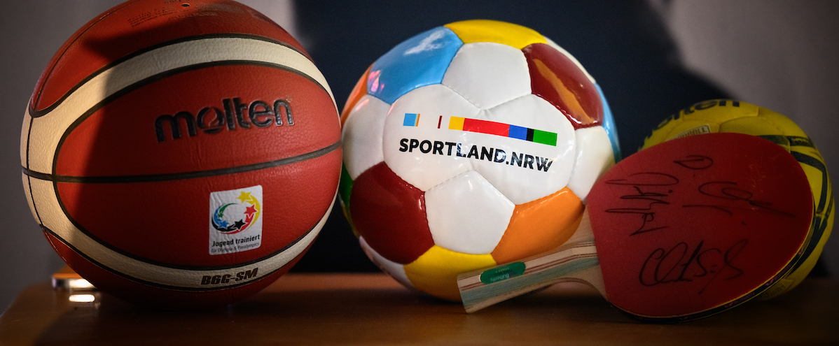 Ball mit Sportland.NRW-Logo neben Basketball und Tischtennisschläger