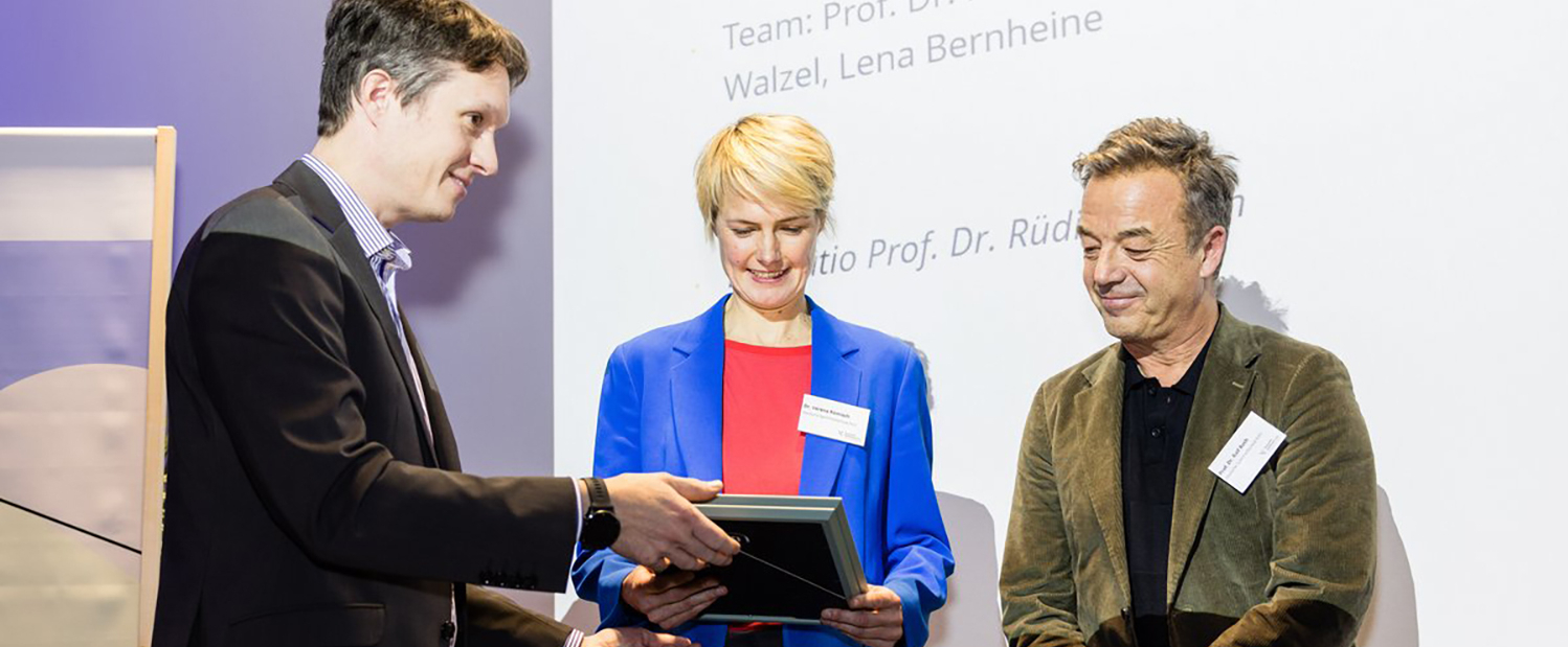 Drei Personen, in der Mitte eine Frau, bei der Übergabe einer Urkunde