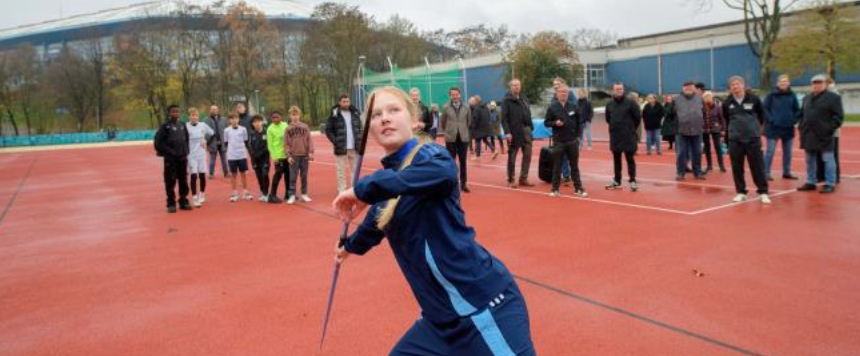 Junge Frau wirft einen Speer, hinter ihr stehen Personen und schauen zu