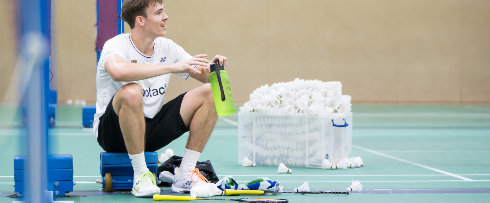 Linus sitzt mit angezogenen Knien in der Badmintonhalle am Netz