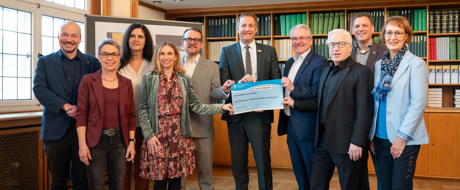 Gruppe von Personen in einem Raum mit Holzfußboden, ein Mann in der Mitte hält einen Bescheid in der Hand