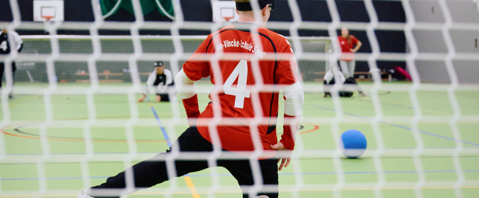 Handballer in rot-schwarzem Dress durch das Tornetz hindurch fotografiert
