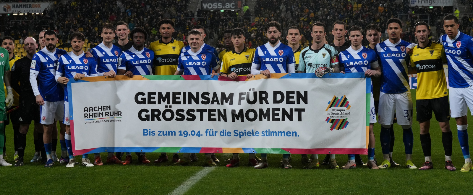 Zwei Fußballmannschaften auf dem Rasen im Stadion halten das Banner der Bewerbung KölnRheinRuhr