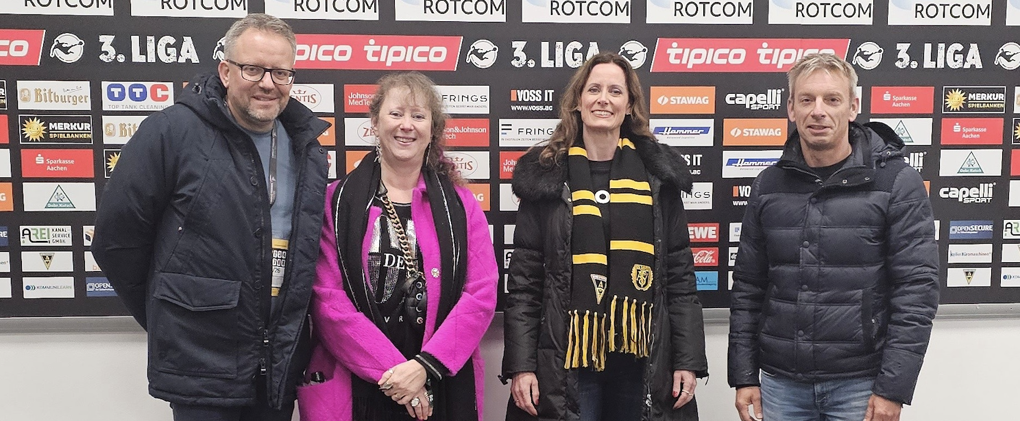 Staatssekretärin Milz neben einer Frau und zwei Männern vor einer Sponsorenwand bei Alemannia Aachen