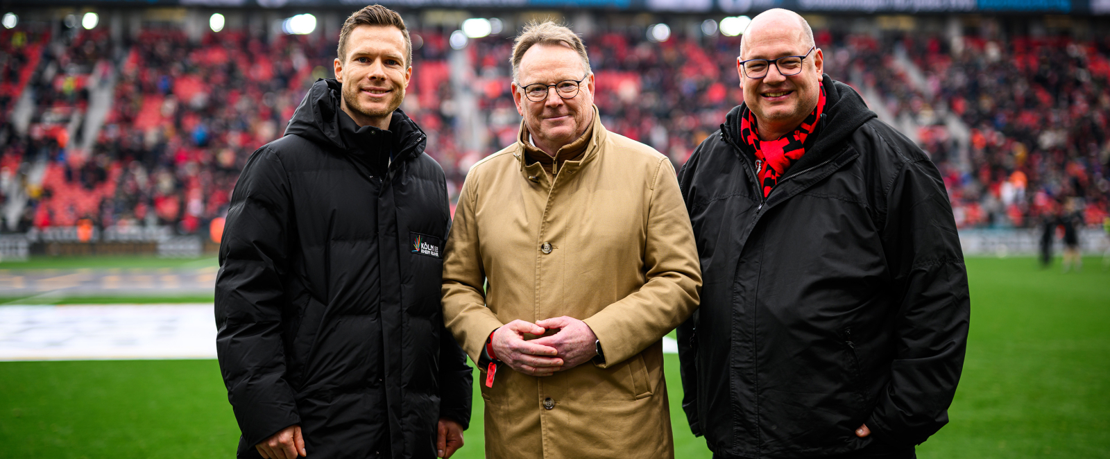 Drei Männer auf dem Rasen in einem Stadion machen Werbung für die Bewerbung KölnRheinRuhr
