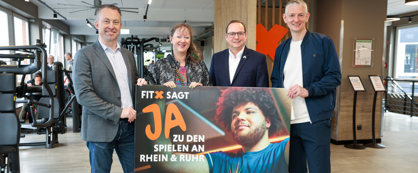 Andrea Milz mit drei Männern in einem Fitnessstudio, sie halten ein Banner der Bewerbung KölnRheinRuhr
