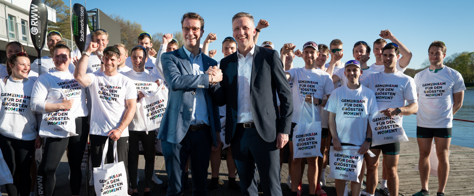 Hendrik Wüst umgeben von Ruderern in weißen Shirts mit der Aufschrift "Gemeinsam für den großen Moment"