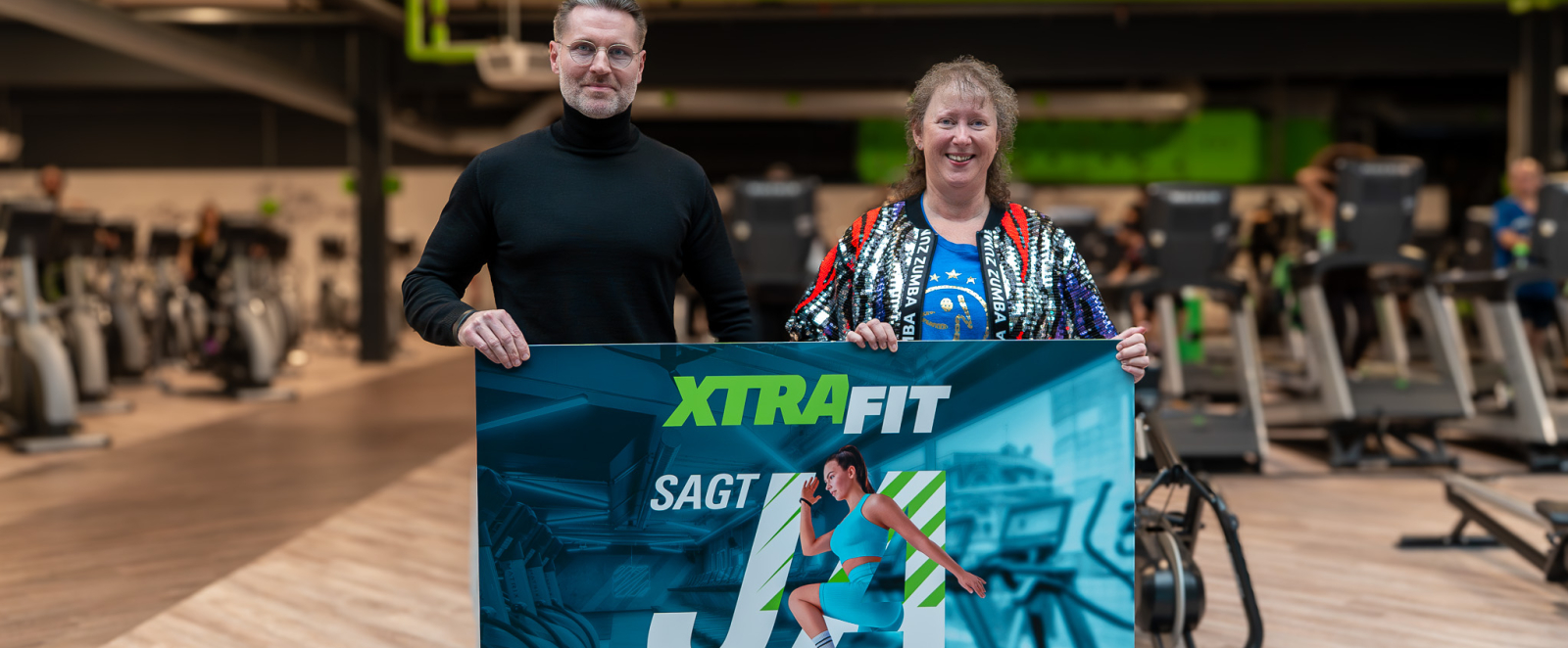 Andrea Milz neben einem Mann in einem Fitnessstudio, beide halten ein Banner zur KRR-Kampagne