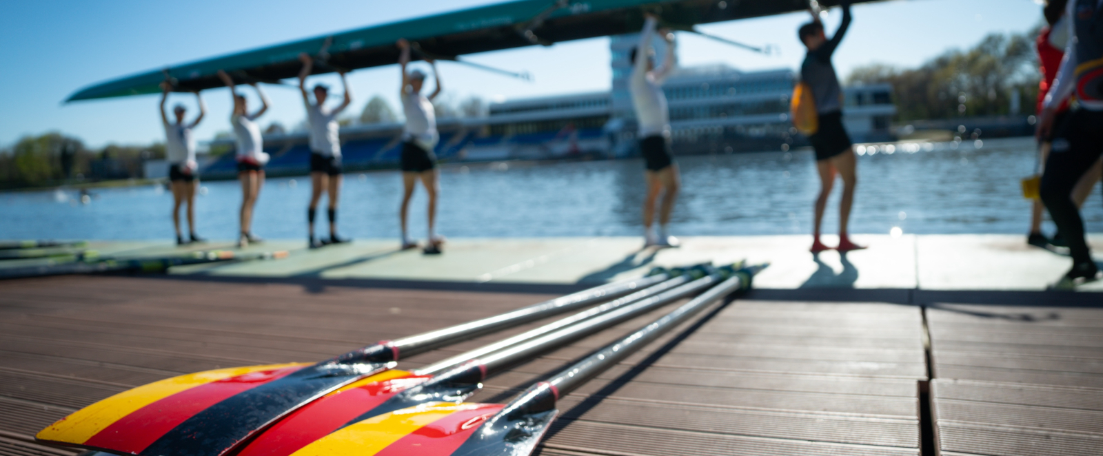 Ruderer mit dem Boot über ihrem Kopf an einer Regattastrecke, im Vordergrund liegen Paddel in Deutschland-Farben
