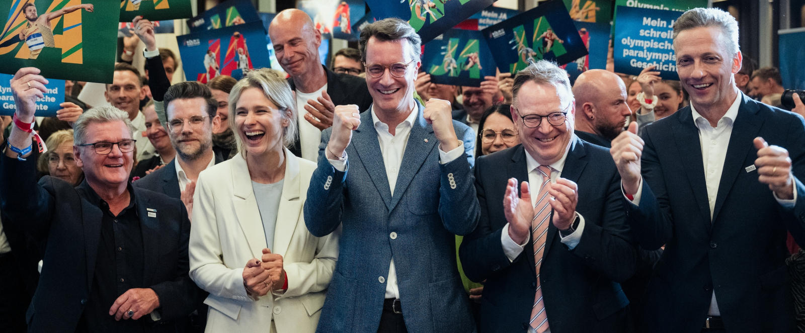 Ministerpräsident Wüst sowie weitere Gesichter aus Politik, Sport, Medien und Wirtschaft jubeln über das Zwischenergebnis des Ratsbürgerentscheides