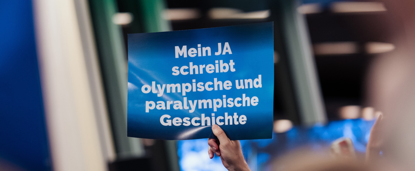 Eine Hand hält ein blaues Schild hoch mit der Aufschrift "Mein JA schreibt olympische und paralympische Geschichte"