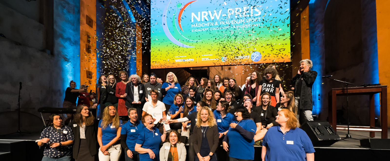 Bühne mit vielen Frauen und Mädchen, dahinter ein Bildschirm mit der Aufschrift "NRW-Preis"