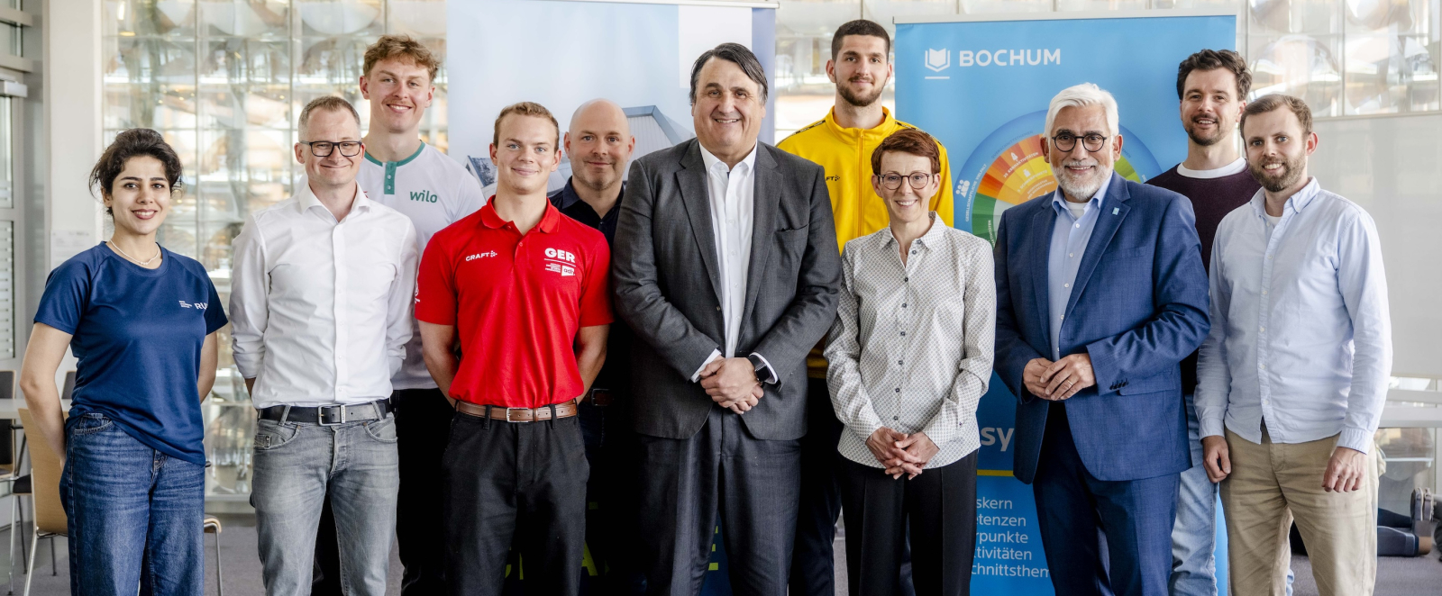Elf Personen in einem Halbkreis vor zwei Bannern und einer Wand aus Glasbausteinen