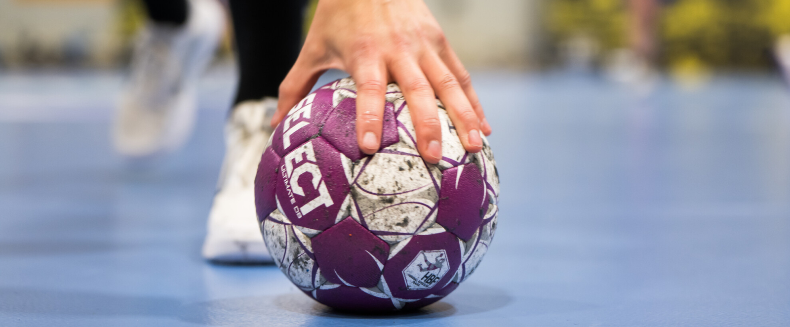 Hand auf einem lila-weißen Handball auf einem blauen Hallenboden