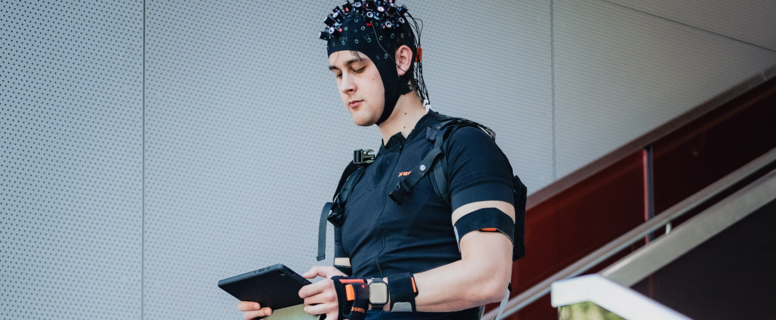 Junger Athlet in blauer Sportkleidung mit Messgeräten am Körper hält ein Tablet in der Hand und geht eine Treppe hinunter