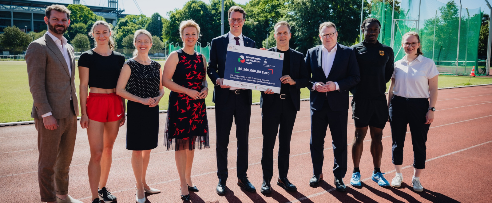 Ministerpräsident Wüst umgeben von acht Personen auf einer Tartanbahn in einem Stadion, er hält ein Schild