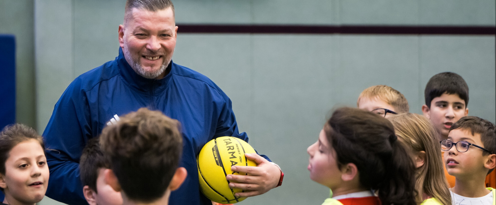 Mann in blauer Sportkleidung hält einen gelben Ball, er ist umgeben von Jungen und Mädchen
