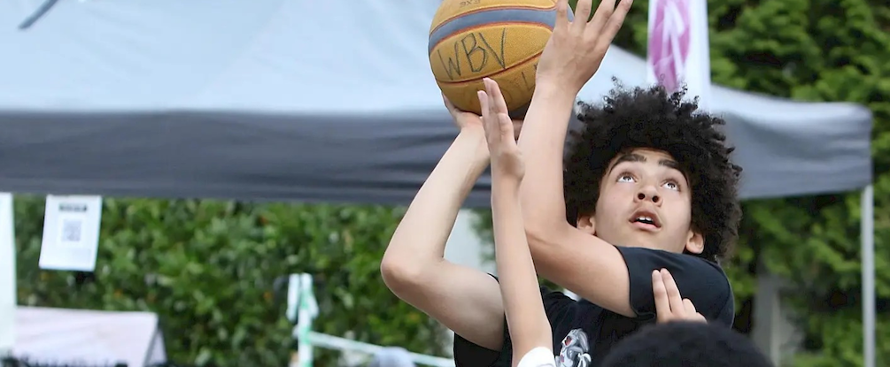 Ein junger Basketballspieler in einem schwarzen Trikot versucht, einen Wurf zu erzielen, während er bei einem Spiel unter freiem Himmel unter einem Vordach eng von Gegnern gedeckt wird.