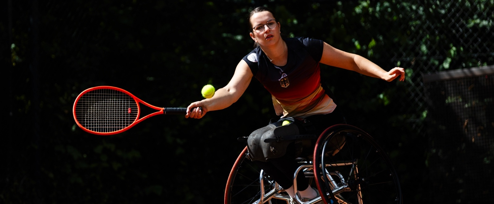 Rollstuhltennisspielerin vor einer dunklen, grünen Hecke