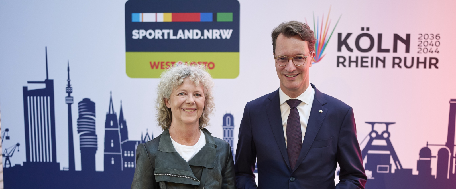 Hendrik Wüst steht rechts von einer Frau vor einer Wand mit dem Logo "Sportland.NRW" und "KölnRheinRuhr"