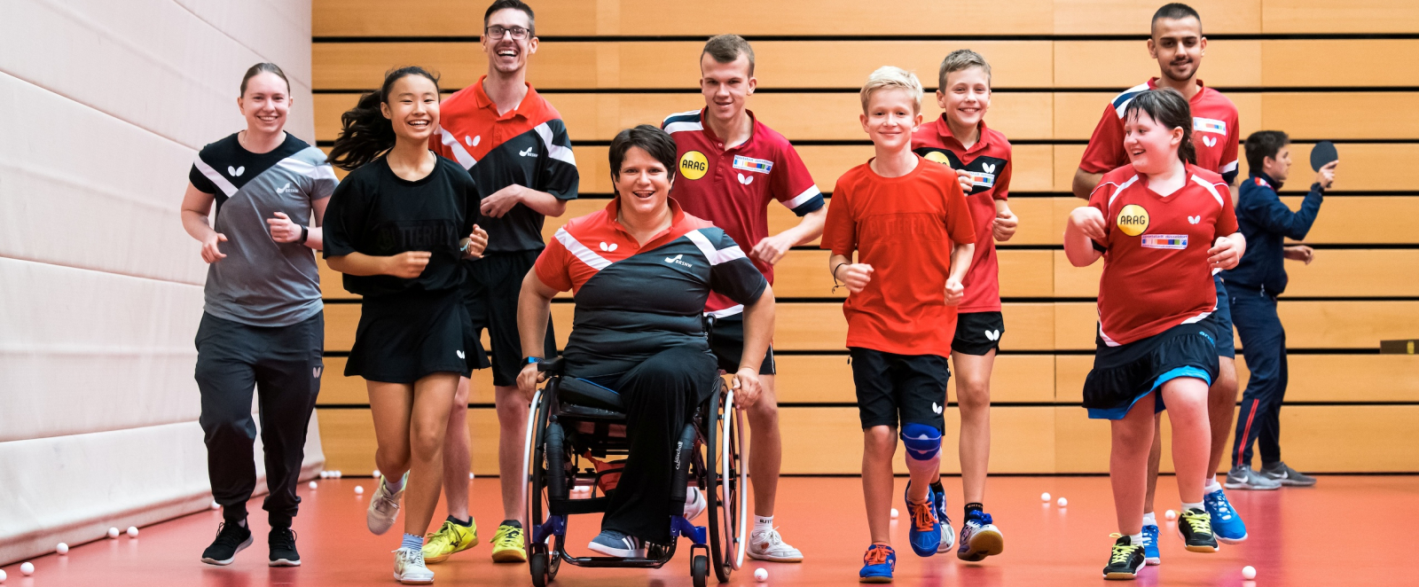 Gruppe von Sportlerinnen und Sportlern, eine davon in einem Rollstuhl, in einer Halle mit rotem Boden