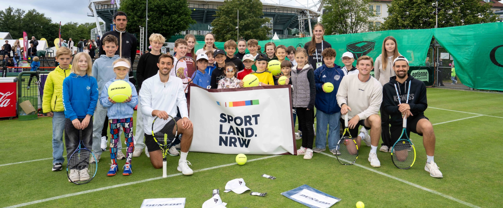 Gruppe von Kindern und jungen Personen halten ein Banner mit "Sportland.NRW", sie sind auf einem Rasen-Tennisplatz