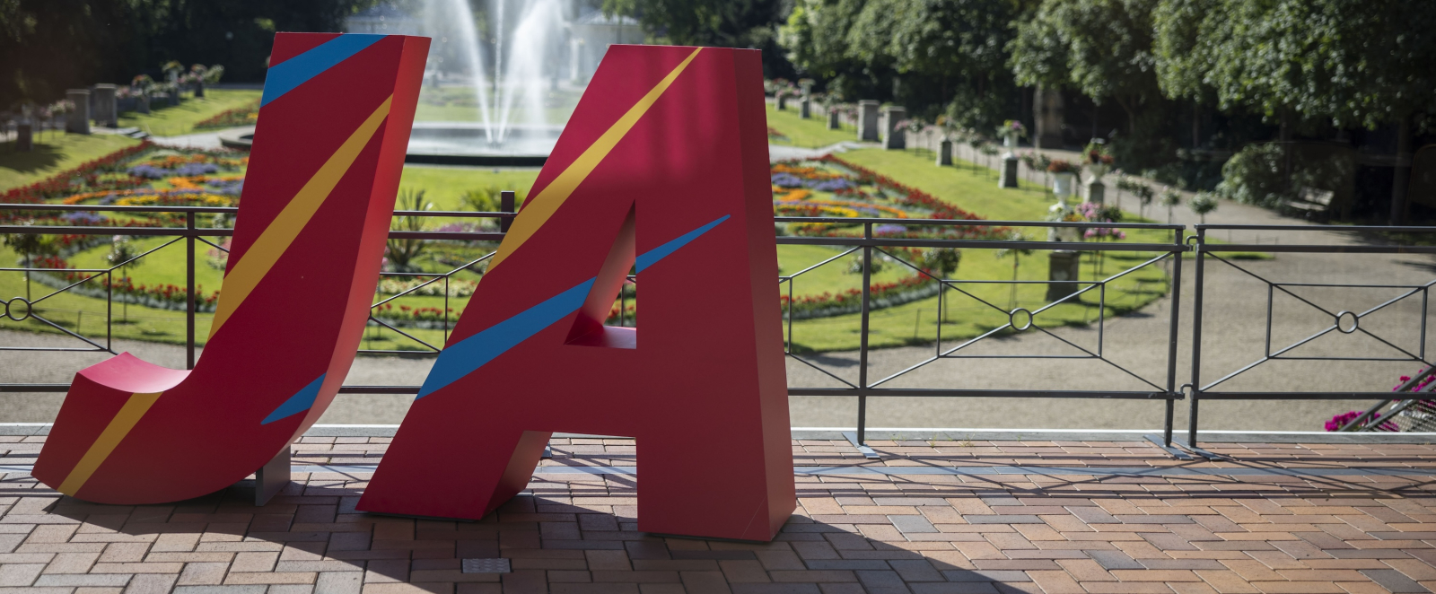 Große rote Buchstaben J und A vor einem Park mit Blumen und Bäumen