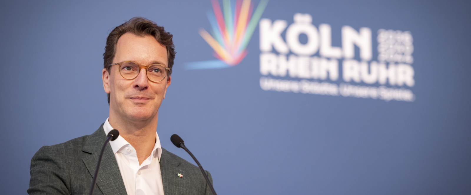 Hendrik Wüst vor einem blauen Banner mit dem Schriftzug KölnRheinRuhr