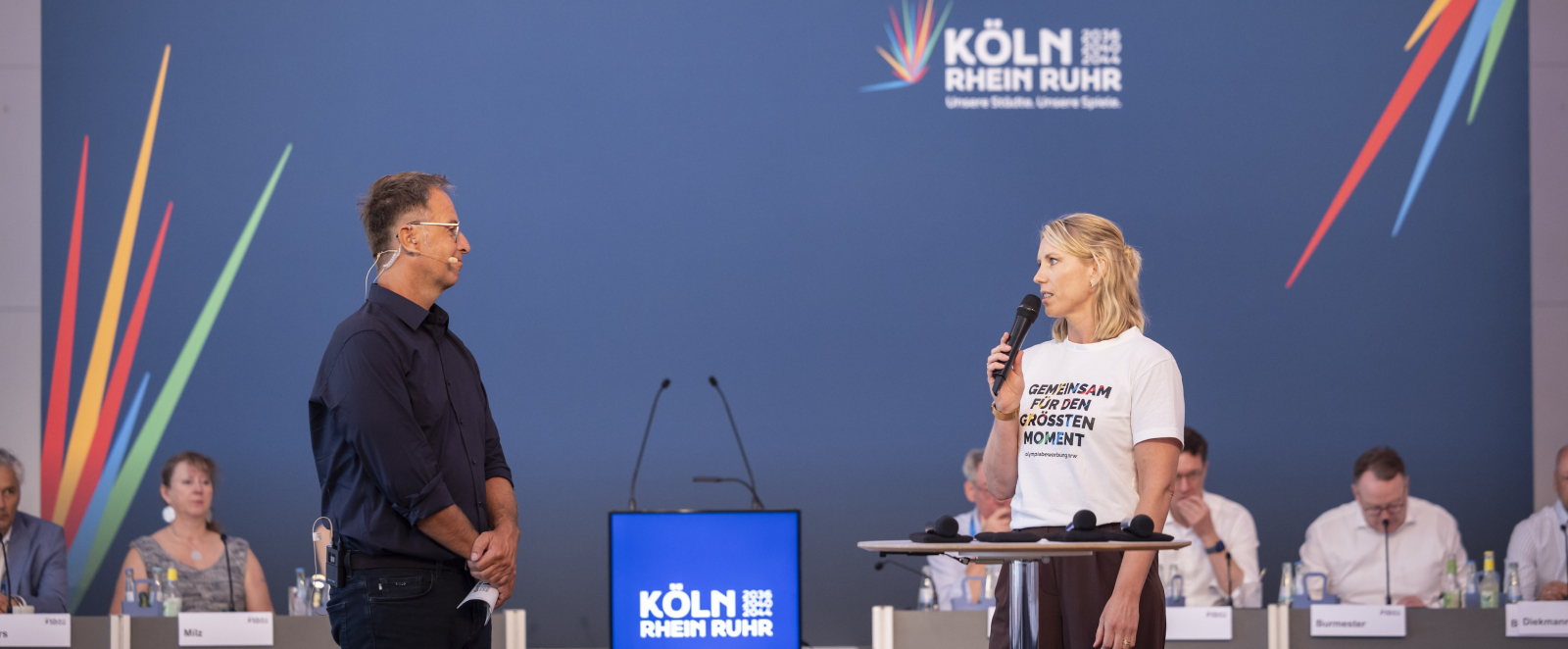 Ein Mann und eine Frau auf einer Bühne vor einem blauen Banner mit dem Schriftzug KölnRheinRuhr