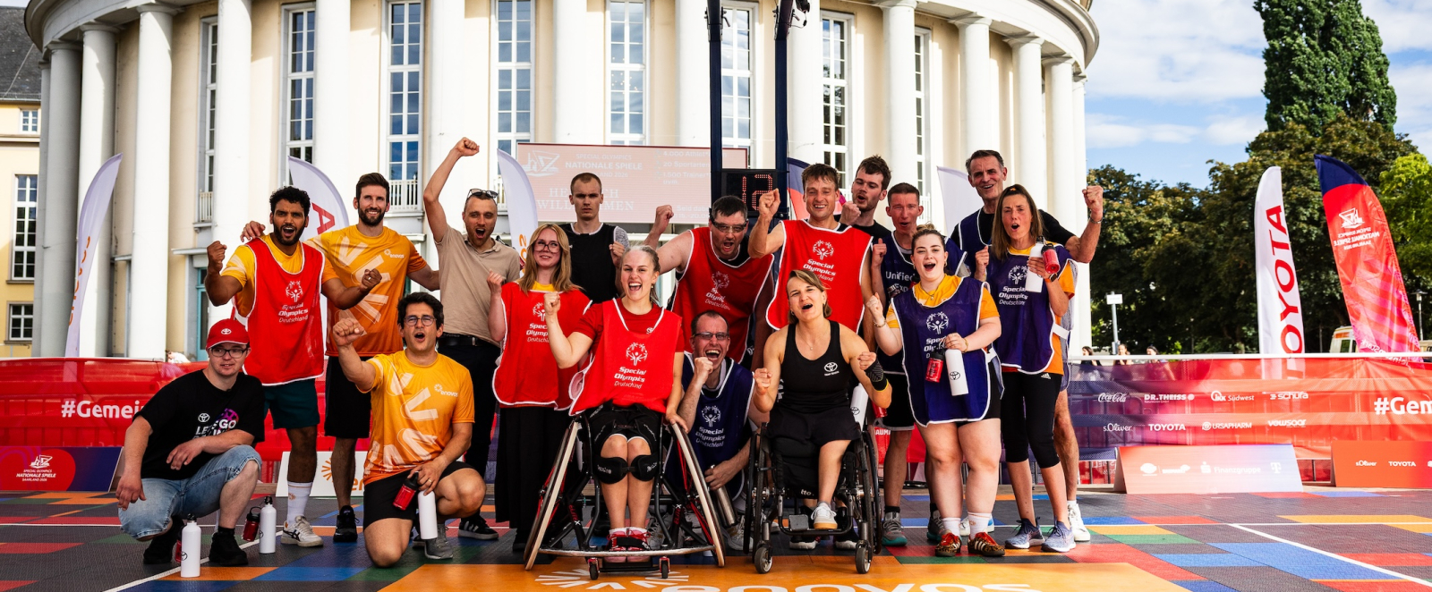 Gruppe von Sportlerinnen und Sportlern, davon eine in einem Rollstuhl auf einem 3x3-Basketballfeld mit einem weißen Gebäude im Hintergrund  