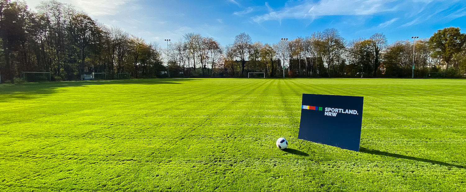 Von Bäumen umgebene grüne Rasenfläche, auf der ein Schild mit Sportland.NRW steht, daneben liegt ein Fußball