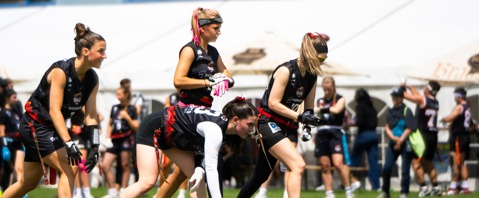 Junge Frauen in schwarzer Sportkleidung spielen Flag Football auf einer Wiese, Personen stehen im Hintergrund