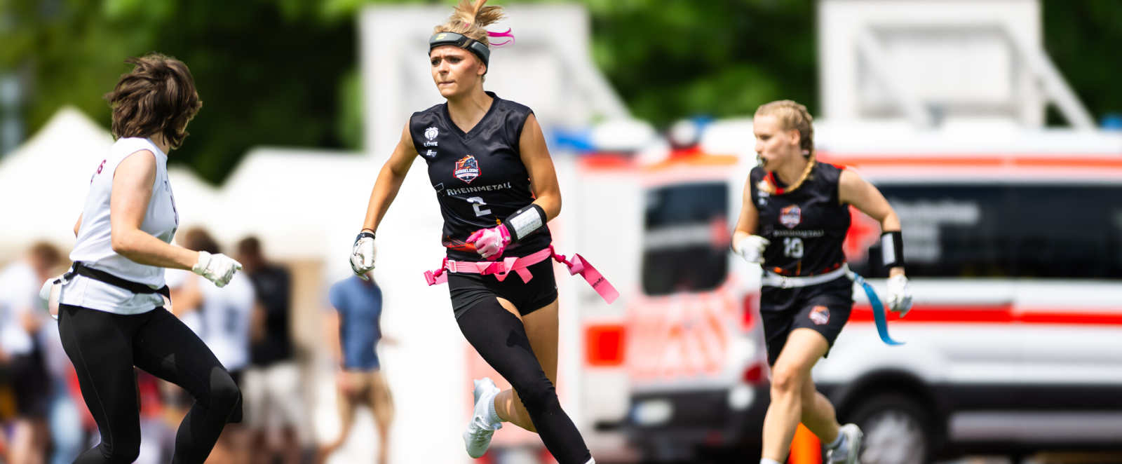 Junge Frau in schwarzer Sportkleidung auf einer Wiese mit Fahrzeugen im Hintergrund spielt Flag Football