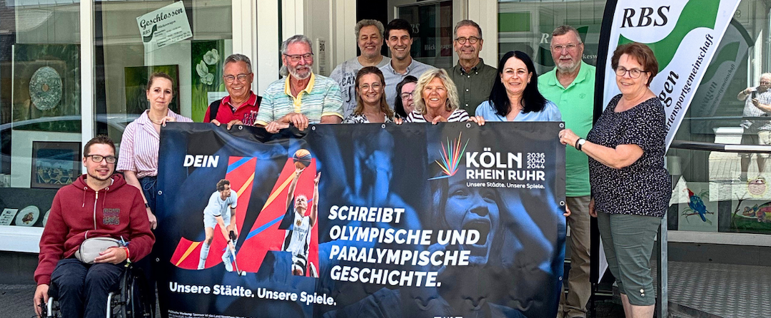 gruppe von Personen, davon einer in einem Rollstuhl, vor einem Gebäude. Sie halten ein Banner der Berwerbung KölnRheinRuhr