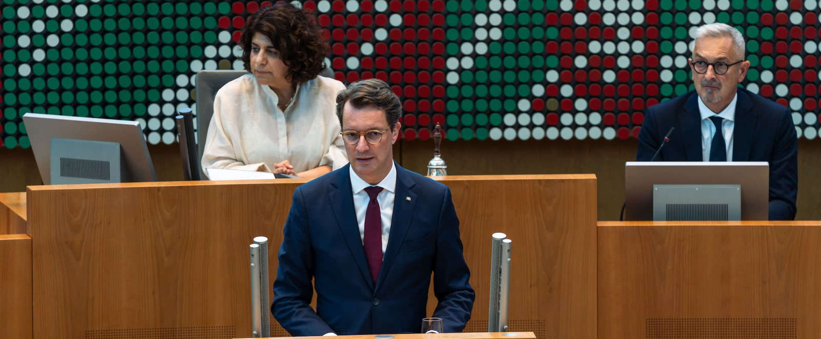 Ministerpräsident Wüst bei einer Rede im Landtag, hinter ihm zwei Personen