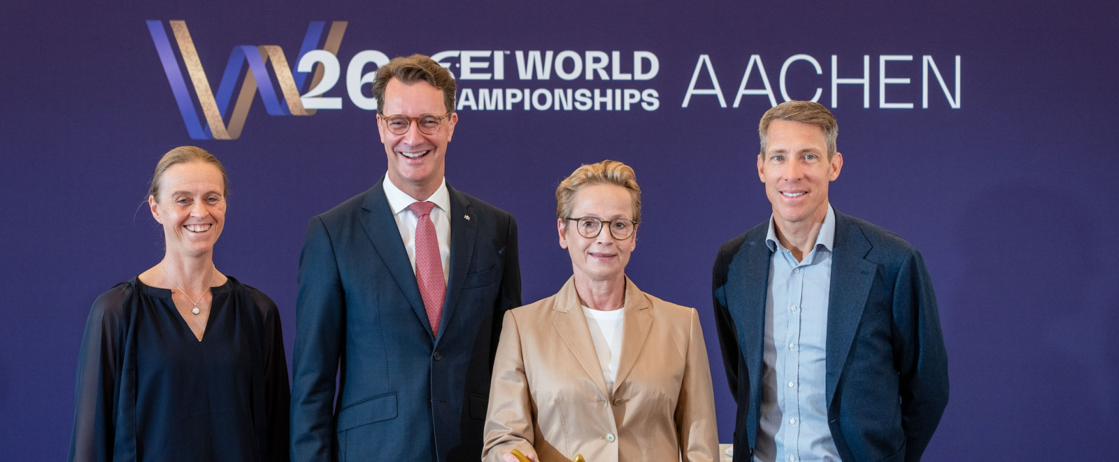 Ministerpräsident Wüst mit drei Pferdesport-Athletinnen und -Athleten vor einem Banner der Reit-WM in Aachen