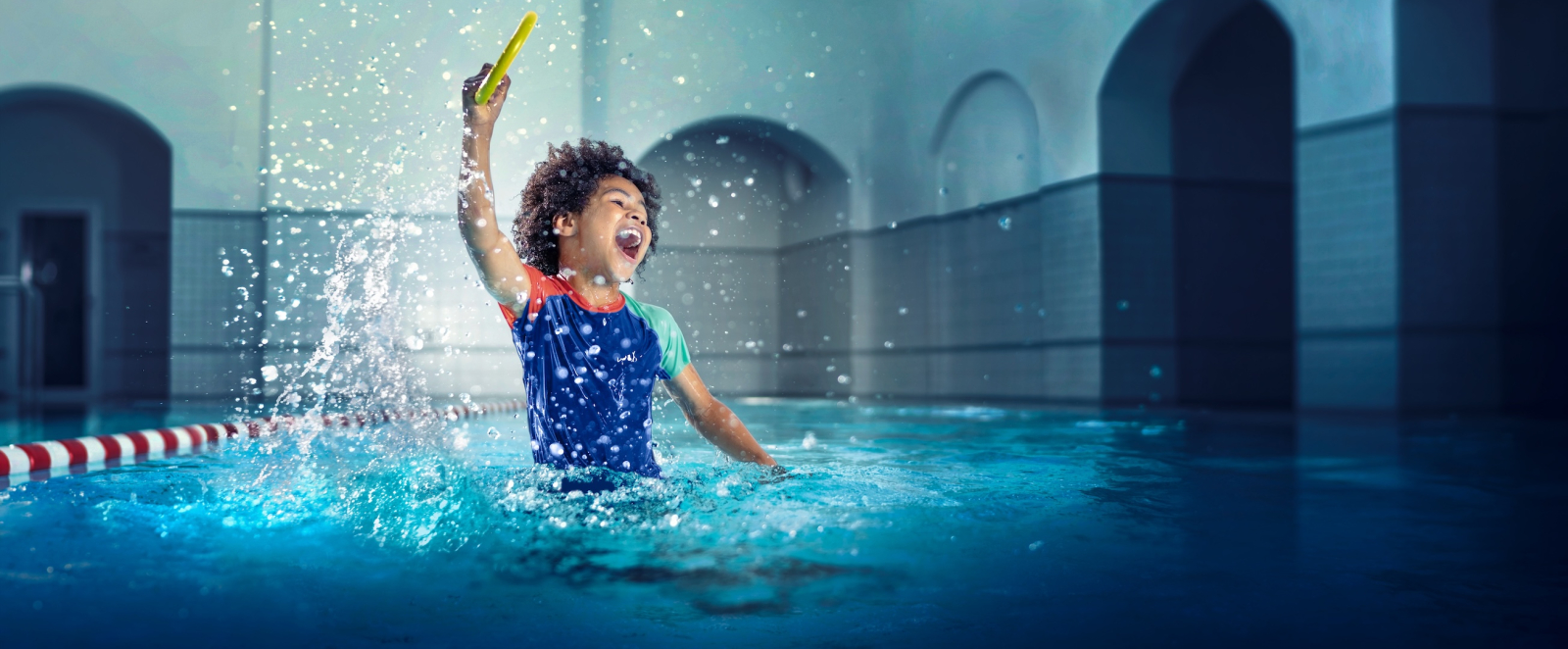 Junge springt hoch in einem Schwimmbecken, im Hintergrund sind Rundbögen zu sehen