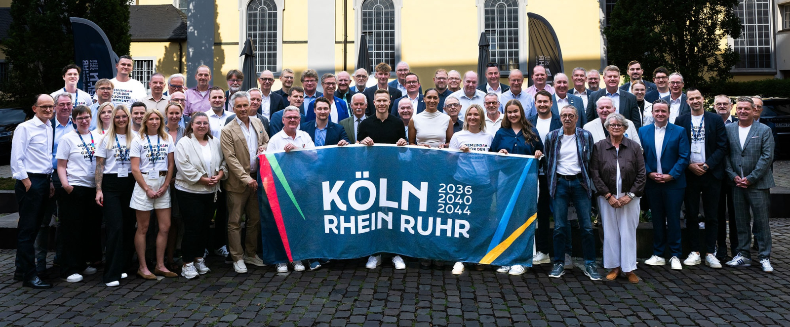 Gruppe von Menschen vor einem beigefarbenen Altbau, sie halten ein Banner der Bewerbung KRR